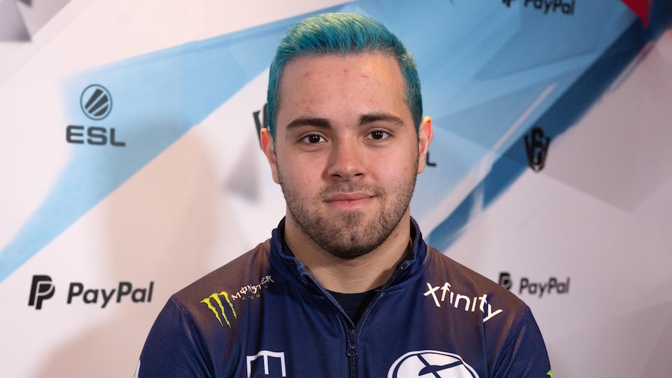 Un jeune homme aux cheveux bleus courts. Il porte une veste bleu marine sur laquelle le nom et le logo de son équipe sont imprimés, ainsi que de nombreux logos de commanditaires.