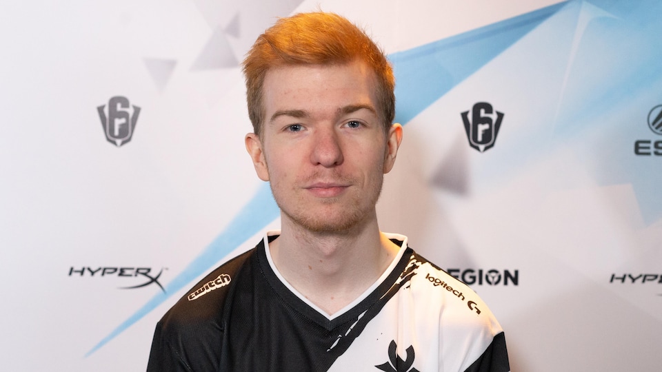 Une photo de Pengu, un jeune homme aux cheveux roux courts. Il porte un t-shirt noir et blanc sur lequel le logo de son équipe et ceux de ses commanditaires sont imprimés.